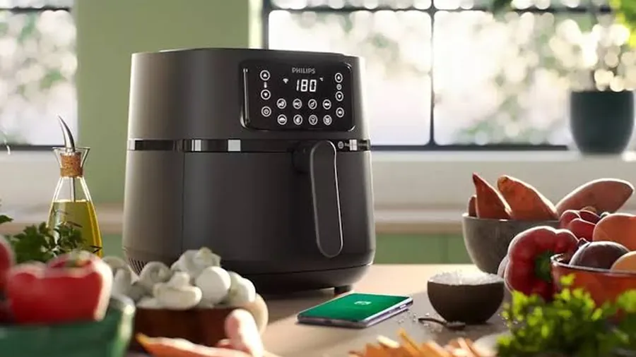 Air fryer noir à écran tactile posé devant une fenêtre, entouré de légumes frais et de fruits dans une cuisine lumineuse