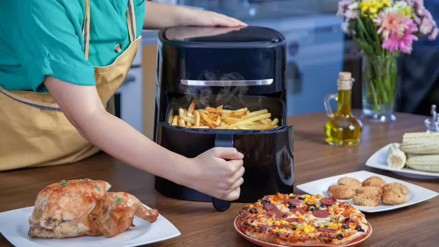 Personne retirant le panier d'un air fryer rempli de frites, entourée d'un poulet rôti, d'une pizza et de beignets servis à table