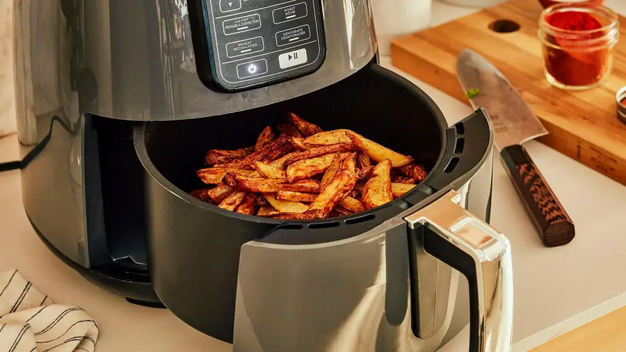Panier d'air fryer contenant des quartiers de pommes de terre épicés répartis en une couche, près d'un couteau et d'une planche à découper