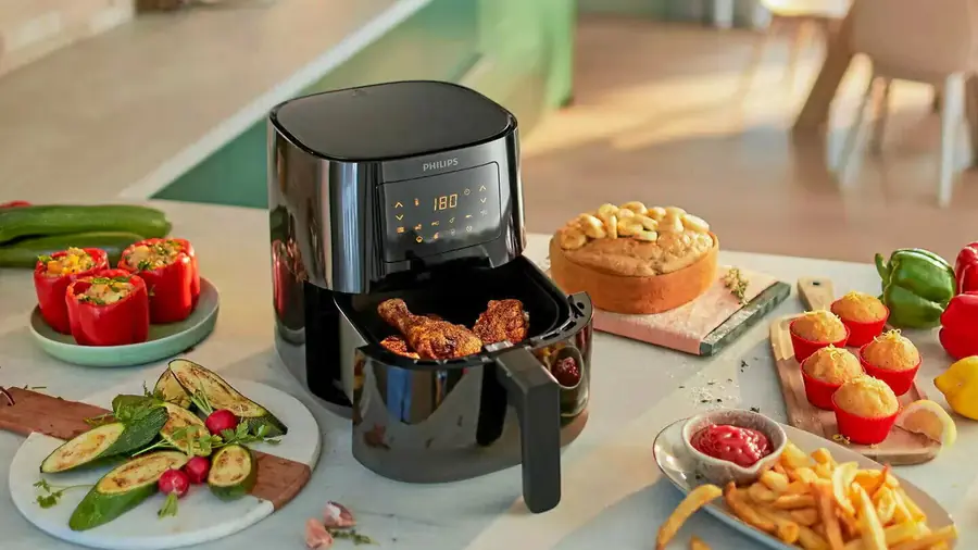 Air fryer à panier ouvert installé sur un plan de travail clair, entouré de légumes farcis, de frites et de pâtisseries