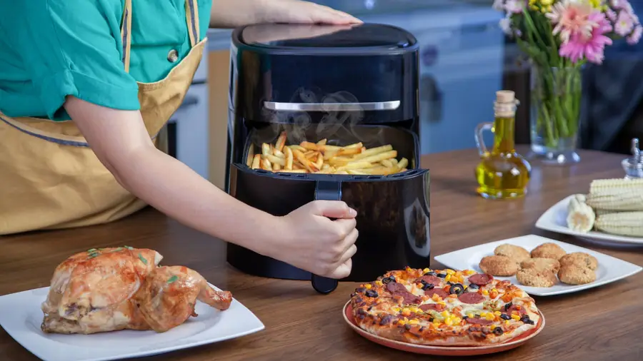 Personne retirant un panier d'air fryer chargé de frites, entourée d'un poulet rôti, d'une pizza et de beignets servis sur la table