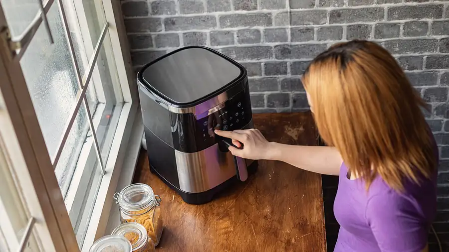 Personne réglant le panneau de commande tactile d'un air fryer posé sur une table en bois près d'une fenêtre